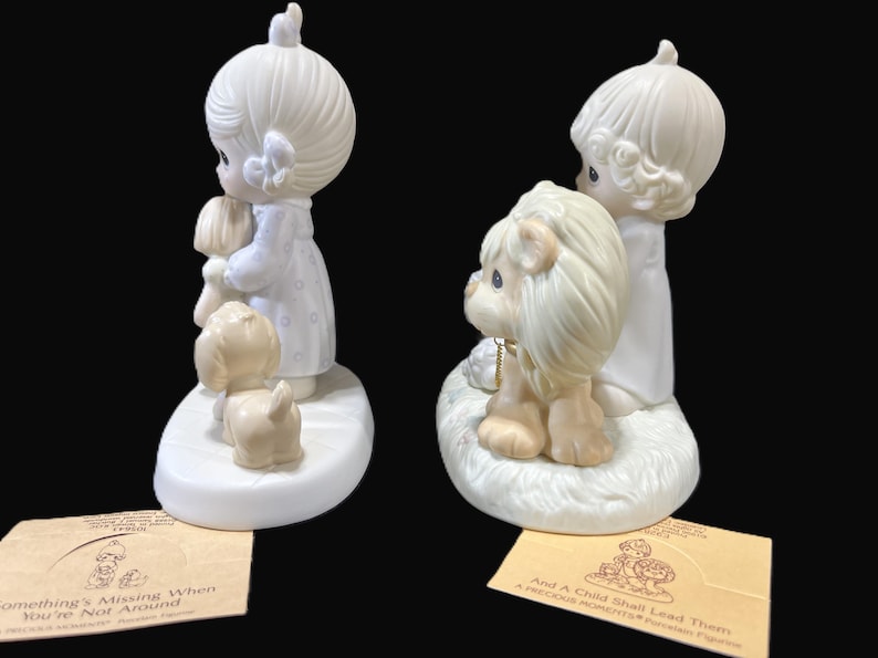 Puede incluir: Dos figuritas de porcelana de ni&ntilde;os, una con un perro y la otra con un le&oacute;n. Las figuritas son blancas con un ligero tono amarillo. La ni&ntilde;a con el perro lleva un perro peque&ntilde;o en brazos. El ni&ntilde;o con el le&oacute;n est&aacute; de pie junto a un le&oacute;n. Las bases de las figuritas son blancas y tienen un ligero tono amarillo. Las bases tienen texto en ellas. El texto en la base de la ni&ntilde;a con el perro dice "Something's Missing When You're Not Around". El texto en la base del ni&ntilde;o con el le&oacute;n dice "And A Child Shall Lead Them A Precious Moments Figurine".