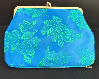 May include: A blue clutch with a gold clasp and frame. The clutch features a repeating pattern of green leaves and floral designs. The bag has a rectangular shape with a rounded top and a zipper closure.