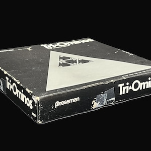 May include: A black box game called "Tri-Ominos" by Pressman. The box has a silver triangle with smaller triangles inside. The game is a classic tile-matching game.