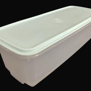 May include: A rectangular, translucent plastic food storage container with a matching lid. The lid has a recessed centre with a circular design and a raised edge. The container is light green and sits against a black background.