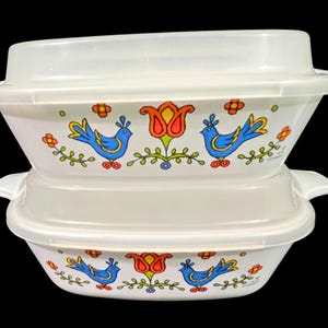 May include: Two white rectangular glass baking dishes stacked vertically, each featuring a translucent plastic lid and side handles. The exterior of the dishes displays a colorful pattern of blue birds, orange tulips, and small green floral accents.
