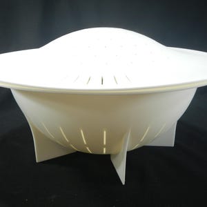 May include: White plastic colander with a removable lid. The colander has a wide base with four legs and a bowl-shaped design. The lid has a flat top and a slightly raised center.