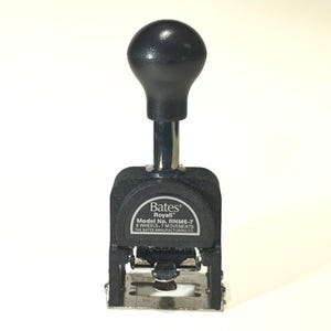 May include: A black Bates Royall numbering stamp with a rounded black handle and a silver metal base. The stamp has the model number RNM6-7 and the text "6 WHEELS, 7 MOVEMENTS". The Bates Manufacturing Co. is also printed on the stamp.