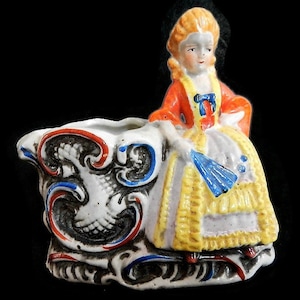May include: A porcelain figurine of a woman in a red, white, and yellow dress sitting on a decorative planter with blue and red accents.