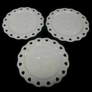 May include: Set of three white ceramic plates with a heart-shaped scalloped edge. Each plate features a raised design of a floral motif.