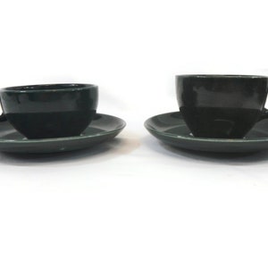 May include: Two dark green ceramic teacups and saucers. The cups have a rounded shape and a small handle. The saucers are slightly wider than the cups and have a smooth, flat surface.
