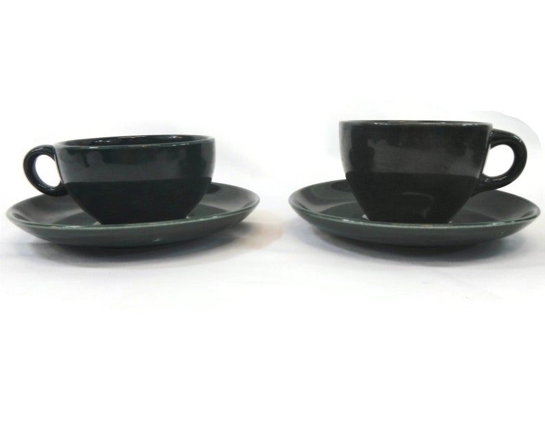 May include: Two dark green ceramic teacups and saucers. The cups have a rounded shape and a small handle. The saucers are slightly wider than the cups and have a smooth, flat surface.