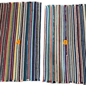 May include: Two rectangular woven rag rugs featuring a dense, vertical striped pattern in a vibrant array of colors including red, blue, yellow, green, black, and white. Each rug is marked with a distinct orange letter, A or B, on its surface.