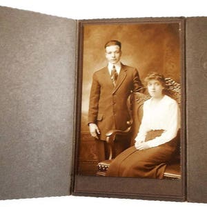 May include: A black and white vintage photograph of a couple posing for a portrait. The man is wearing a suit and tie, and the woman is wearing a white dress. They are standing in front of a chair.