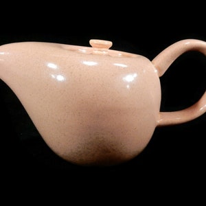May include: A light pink ceramic teapot with a rounded body and a curved spout. The teapot has a smooth, glossy finish and a simple, elegant design.