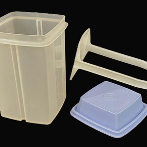 May include: A translucent, square-shaped plastic storage container with a matching light blue lid and a separate white plastic insert. The container features vertical ridges along its sides, while the lid has a flat top and a raised square center.