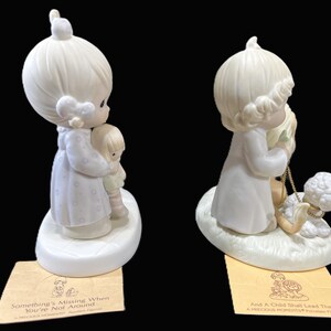 Puede incluir: Dos figuritas de porcelana de personajes de Precious Moments. La figurita de la izquierda representa a una ni&ntilde;a sosteniendo una mu&ntilde;eca. La figurita de la derecha representa a una ni&ntilde;a sosteniendo un cordero. Ambas figuritas tienen una base blanca con un borde dorado. La base de la figurita de la izquierda tiene el texto "Something's Missing When You're Not Around". La base de la figurita de la derecha tiene el texto "And A Child Shall Lead Them".
