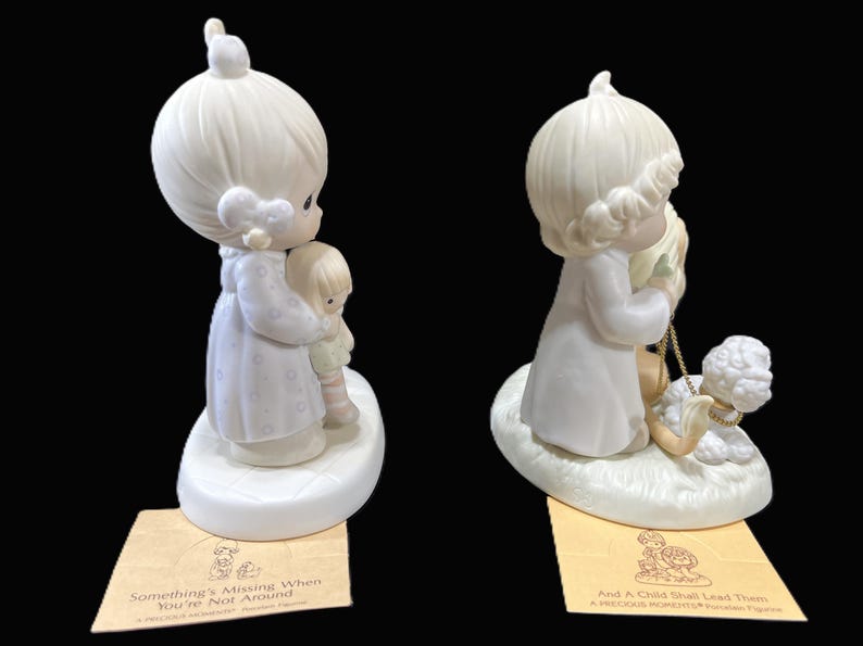 Puede incluir: Dos figuritas de porcelana de personajes de Precious Moments. La figurita de la izquierda representa a una ni&ntilde;a sosteniendo una mu&ntilde;eca. La figurita de la derecha representa a una ni&ntilde;a sosteniendo un cordero. Ambas figuritas tienen una base blanca con un borde dorado. La base de la figurita de la izquierda tiene el texto "Something's Missing When You're Not Around". La base de la figurita de la derecha tiene el texto "And A Child Shall Lead Them".