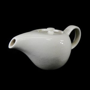 May include: White ceramic teapot with a rounded body and a spout. The teapot has a lid with a small knob on top.