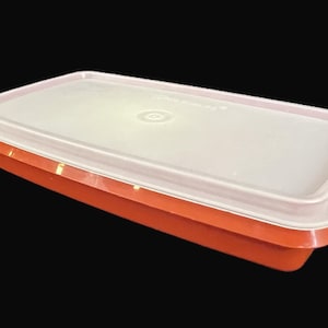 May include: An orange rectangular food storage container with a white lid. The lid has a slightly rounded edge and a circular design in the center. The container is made of plastic and is likely used for storing food items.