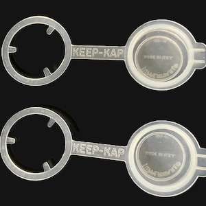 May include: Two translucent, flexible plastic bottle caps featuring a circular ring connected to a solid cap by a flat tab. The text KEEP-KAP is embossed on the tab, while the cap surface displays the text TUPPERWARE and MADE IN U.S.A.