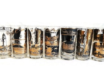 May include: A set of seven clear glass tumblers featuring black and gold printed graphics of various United States landmarks and locations, including Cape Cod, Estes Park, Hyde Park, and San Francisco, with descriptive text for each.