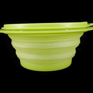 May include: A collapsible, lime green silicone bowl with a matching lid. The bowl has a ridged, accordion-like design, allowing it to expand and contract. The lid sits on top with a small lip for easy removal. The background is black.
