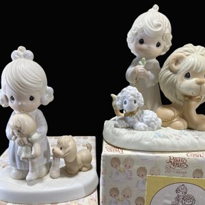 Puede incluir: Dos figuritas de porcelana de personajes de Precious Moments. Una figurita representa a una ni&ntilde;a sosteniendo una mu&ntilde;eca, mientras que la otra figurita representa a una ni&ntilde;a con un cordero y un le&oacute;n. Ambas figuritas est&aacute;n sobre una base blanca.