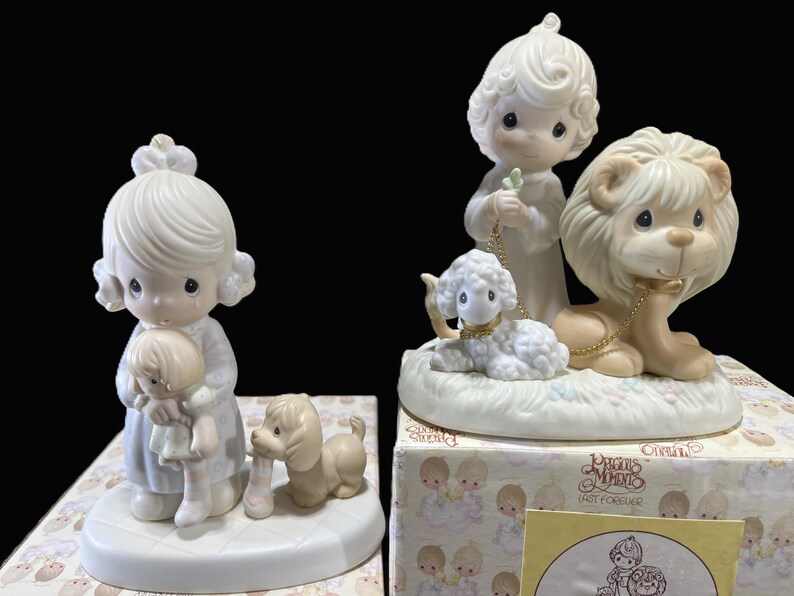 Puede incluir: Dos figuritas de porcelana de personajes de Precious Moments. Una figurita representa a una ni&ntilde;a sosteniendo una mu&ntilde;eca, mientras que la otra figurita representa a una ni&ntilde;a con un cordero y un le&oacute;n. Ambas figuritas est&aacute;n sobre una base blanca.