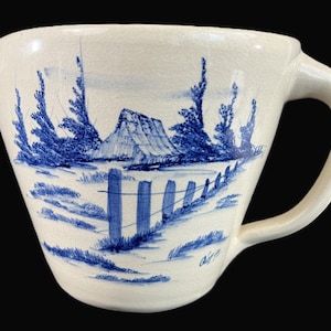 May include: A white ceramic jug with a blue hand-painted design of a mountain scene with trees and a fence. The jug has a wide mouth and a handle.