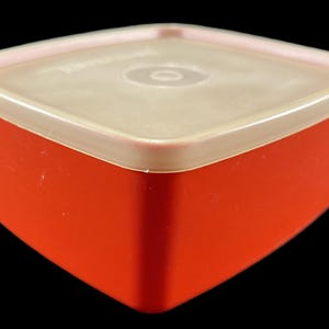 May include: A red square plastic container with a beige lid. The container has a smooth surface and the lid has a slight curve.