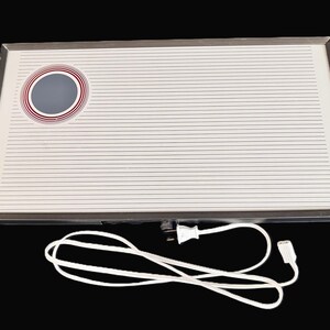 May include: A rectangular electric warming tray with a white surface featuring thin horizontal black lines and a circular graphic with red rings. The tray has a metallic frame, two brown wooden side handles, and a white power cord with a plug.