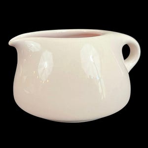 May include: A small, off-white ceramic creamer with a handle and a spout. The creamer has a rounded body and a smooth, glossy finish. The interior is a light pink colour. The creamer is set against a black background.