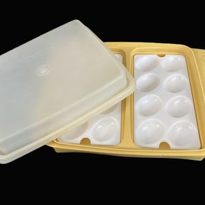 May include: A vintage Tupperware egg container with a yellow base and a translucent lid. The container has two sections, each with oval-shaped compartments to hold eggs. The lid is partially open, revealing the egg holders.