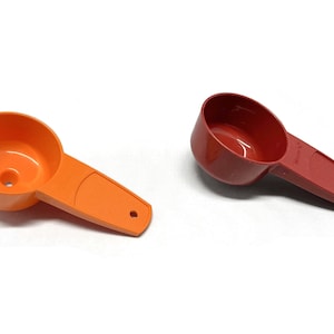 May include: Two plastic measuring cups, one orange and one red. The orange cup has a funnel shape and the red cup has a rounded shape. Both cups have a handle.