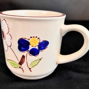 May include: Cream-colored ceramic mug with a brown rim and handle. The mug is decorated with hand-painted flowers in blue, yellow, and white, with green leaves and brown stems. A charming floral design for everyday use.