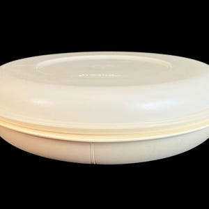 May include: Off-white, vintage Tupperware serving bowl with a matching lid. The bowl has a rounded shape with a slightly raised rim and a fitted lid. The lid and bowl are made of a durable, lightweight plastic. The bowl is designed for food storage and serving.
