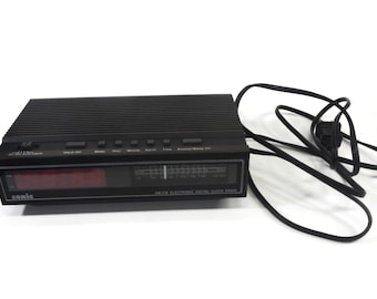 May include: A black Sonic AM/FM electronic digital clock radio with a rectangular housing, a red LED display, and a manual radio tuning dial. The top panel features several control buttons, and a black power cord is attached to the side.