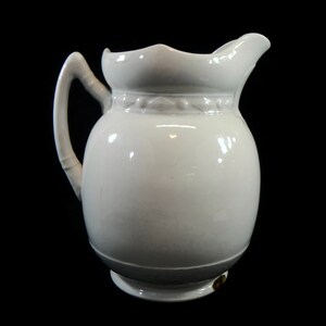 May include: White ceramic pitcher with a rounded body and a scalloped rim. The pitcher has a single handle and a decorative band around the top.