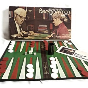 May include: A green and red backgammon board game set with white and red playing pieces, two dice, and a black cup. The box for the game is in the background and features a photo of two people playing backgammon. The box reads "Oswald Jacoby Backgammon".