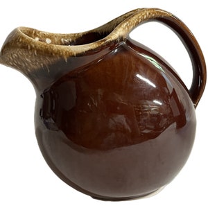 May include: A brown glazed ceramic pitcher with a rounded body and a handle. The pitcher has a light brown glaze on the rim and handle.