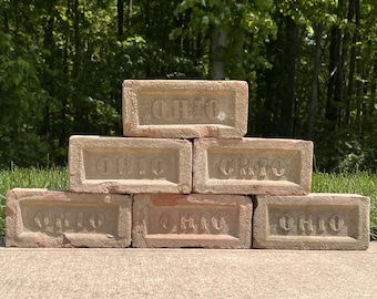 May include: A stack of six rectangular, weathered clay bricks arranged in a pyramid formation. Each brick features the word OHIO embossed in capital letters within a recessed rectangular panel on the front face, showing a rustic, textured surface.