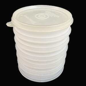 May include: Stack of clear, round plastic food storage containers with lids. The lids are flat with a slightly raised edge and a small tab. The containers are stacked on top of each other, with the lids on top.