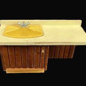 May include: A miniature kitchen sink with a yellow basin and a silver faucet. The countertop is light yellow, and the base is made of brown wood with vertical slats. A small cabinet door is visible.