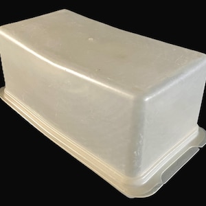 May include: An off-white plastic container, rectangular with a curved top and ridged base. The container has rounded corners and a smooth surface. The base features small, curved cutouts on the sides.