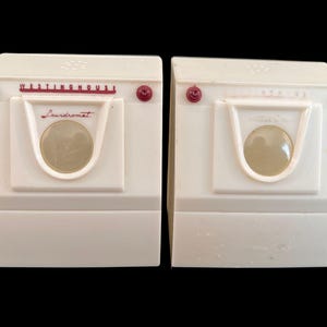 May include: Two vintage white toy washing machines with red accents. Each has a clear porthole and the word "Westinghouse" and "Laundromat" printed on the front. The toys are set against a black background.