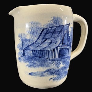 May include: A white ceramic jug with a blue and white design of a barn in a field. The jug has a handle and a spout.