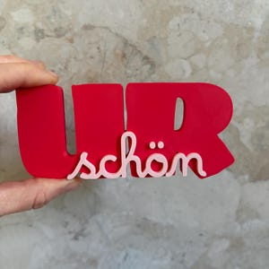 Pode incluir: Uma placa decorativa vermelha brilhante com o texto em negrito UR, com a palavra schön escrita em letra cursiva rosa claro sobreposta na frente. O objeto é construído com um acabamento fosco liso e uma tipografia distinta.