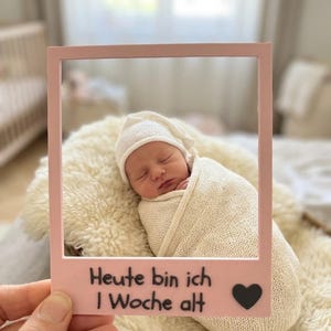 Könnte beinhalten: Ein hellrosa rechteckiger Fotorahmen mit dem aufgedruckten deutschen Text "Heute bin ich 1 Woche alt" und einem kleinen schwarzen Herzsymbol am unteren Rand, gehalten vor einem Neugeborenen in einer cremefarbenen Strickdecke und Mütze.