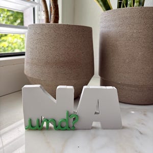 May include: A white decorative sign featuring the bold letters N and A with the green lowercase text und? attached to the front. The object is constructed with a solid, block-like structure and a smooth finish, displaying the German phrase NA und? clearly.