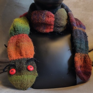 Op de afbeelding: Een kleurrijke gebreide sjaal met een wormdesign. De sjaal is gemaakt van verschillende tinten oranje, groen, bruin en zwart garen. De worm heeft twee grote rode knopen als ogen.
