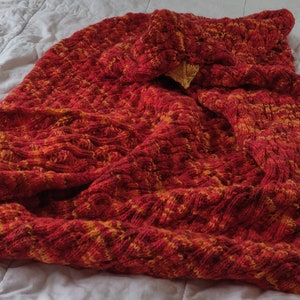 May include: A red and orange knitted blanket with a cable knit pattern. The blanket is folded and draped over a white surface.