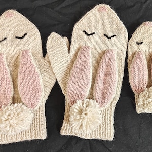 Op de afbeelding: Drie paar gebreide konijnenwantjes in beige en roze. De wantjes hebben een pompon aan de onderkant en zijn ontworpen om eruit te zien als konijnenoren.