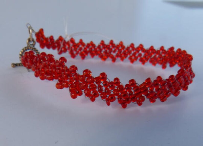 Puede incluir: Una pulsera de cuentas rojas con un cierre decorativo. La pulsera est&aacute; hecha de peque&ntilde;as cuentas rojas redondas ensartadas en un patr&oacute;n en zigzag. El cierre es de color bronce.