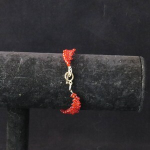 Puede incluir: Una pulsera de cuentas rojas con un cierre plateado. La pulsera est&aacute; hecha de peque&ntilde;as cuentas rojas redondas y se muestra sobre un soporte negro sobre un fondo oscuro. La pulsera mide aproximadamente 18 cm de largo.